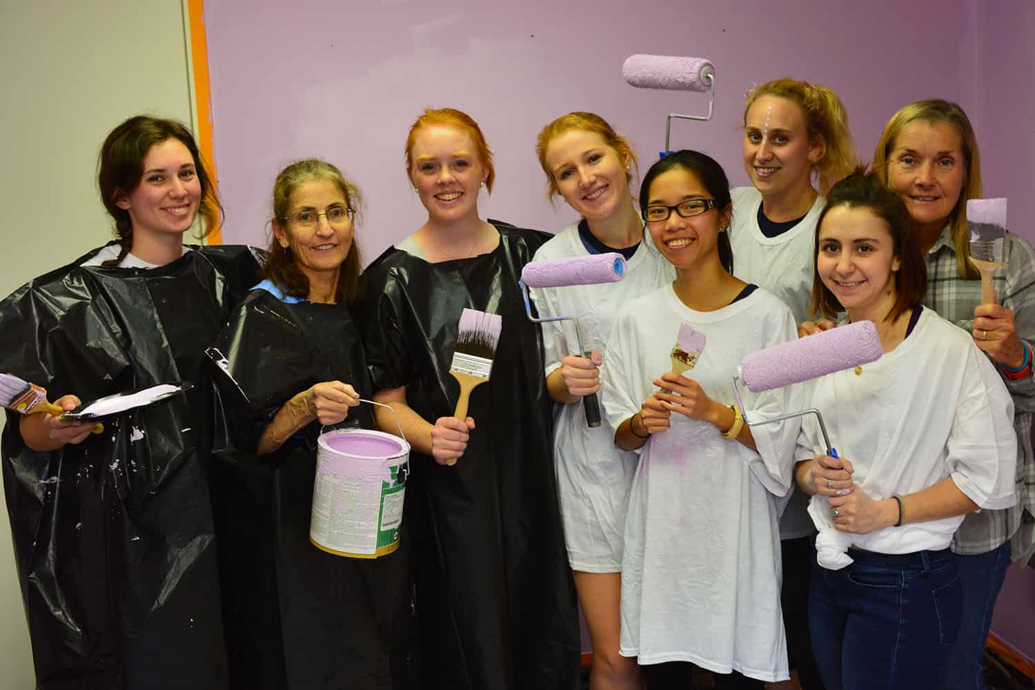 Eight volunteers pose with paint rollers and brushes against the lavender salon wall.