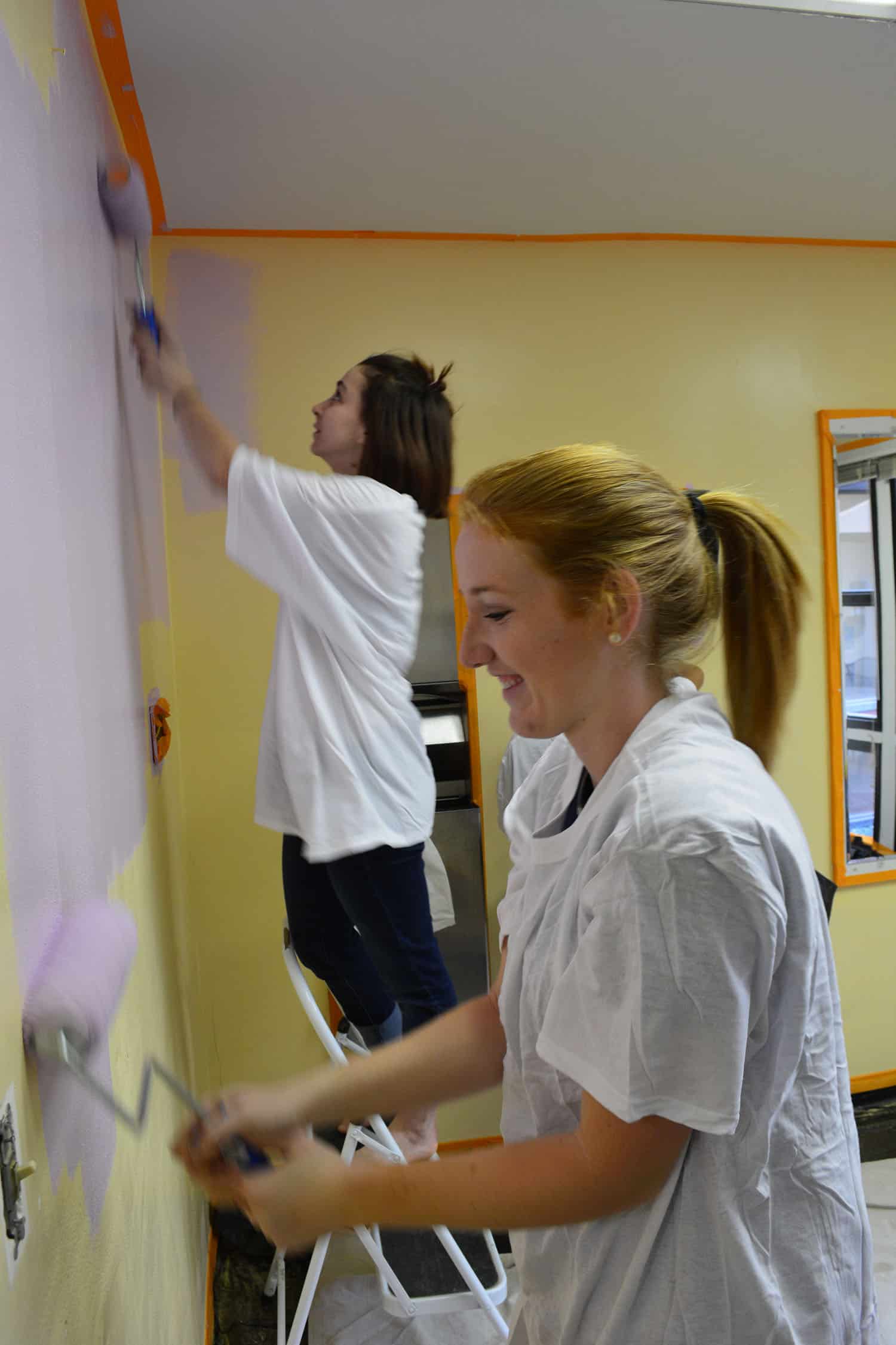 Two volunteers paint the salon wall, one working from a stepladder.