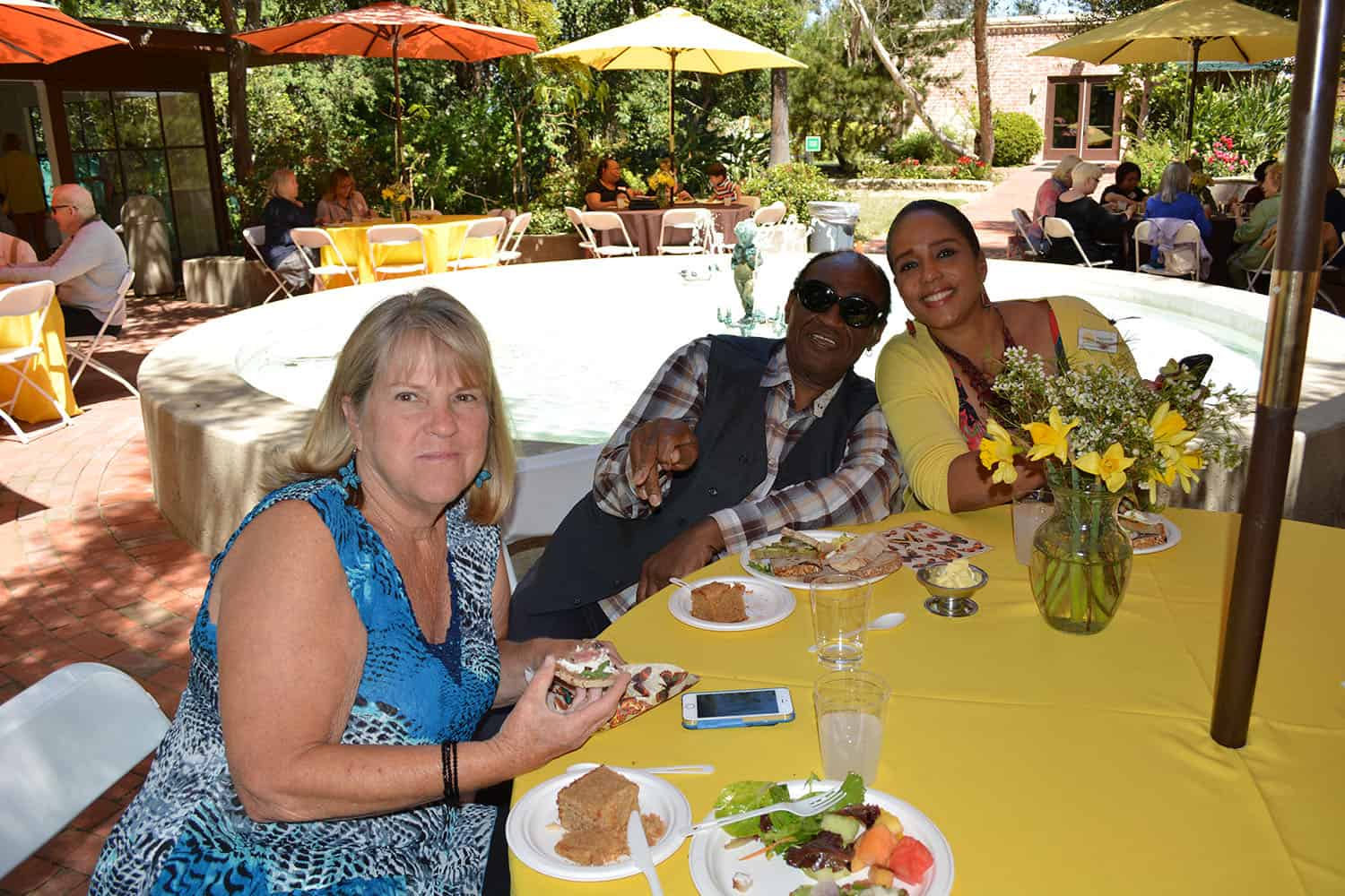 Three attendees smile over lunch at a yellow-covered table beside the fountain.