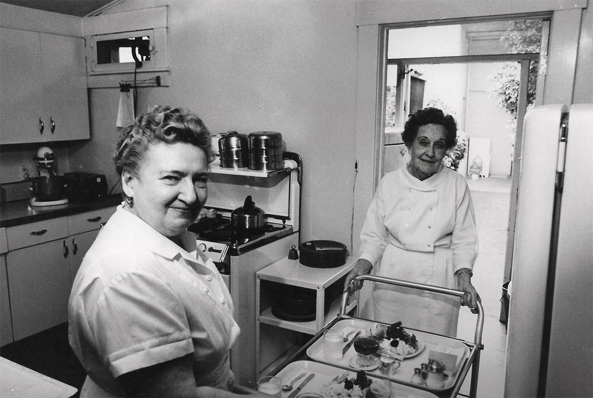 Two women in white uniforms stand with meal trays in a small kitchen.