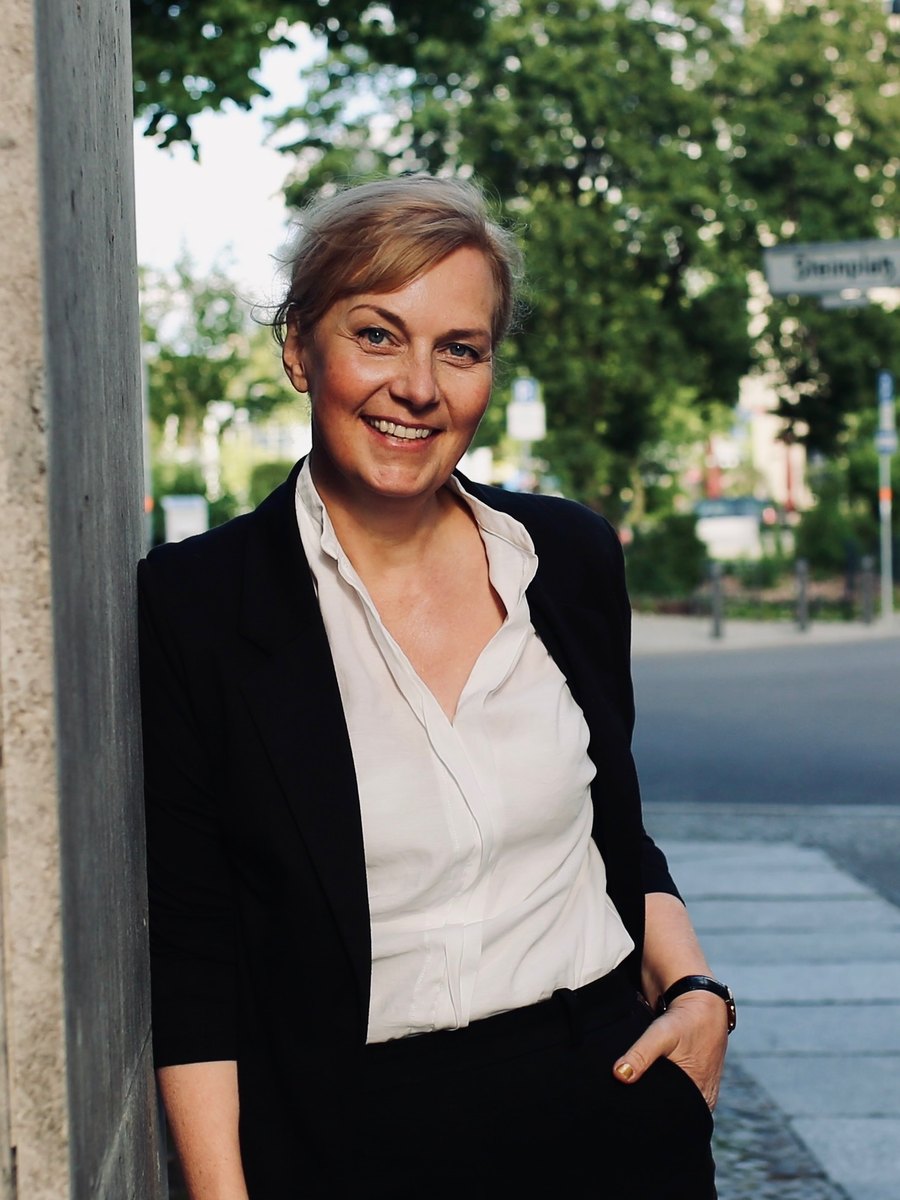 Dr. Annette Kreutziger-Herr, C.S., smiling, leaning against a stone column on a tree-lined street.
